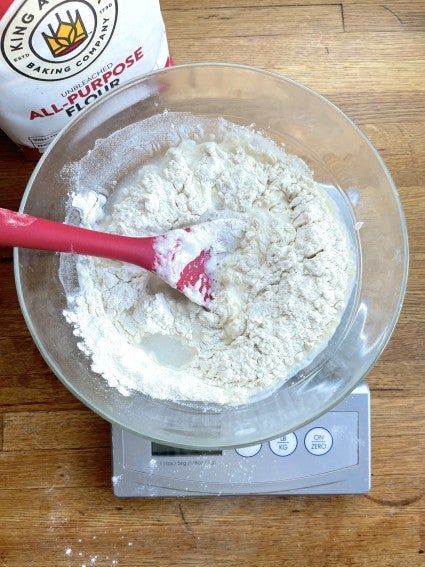 Bowl of sourdough starter on a scale, water and flour being stirred in as part of the feeding process.